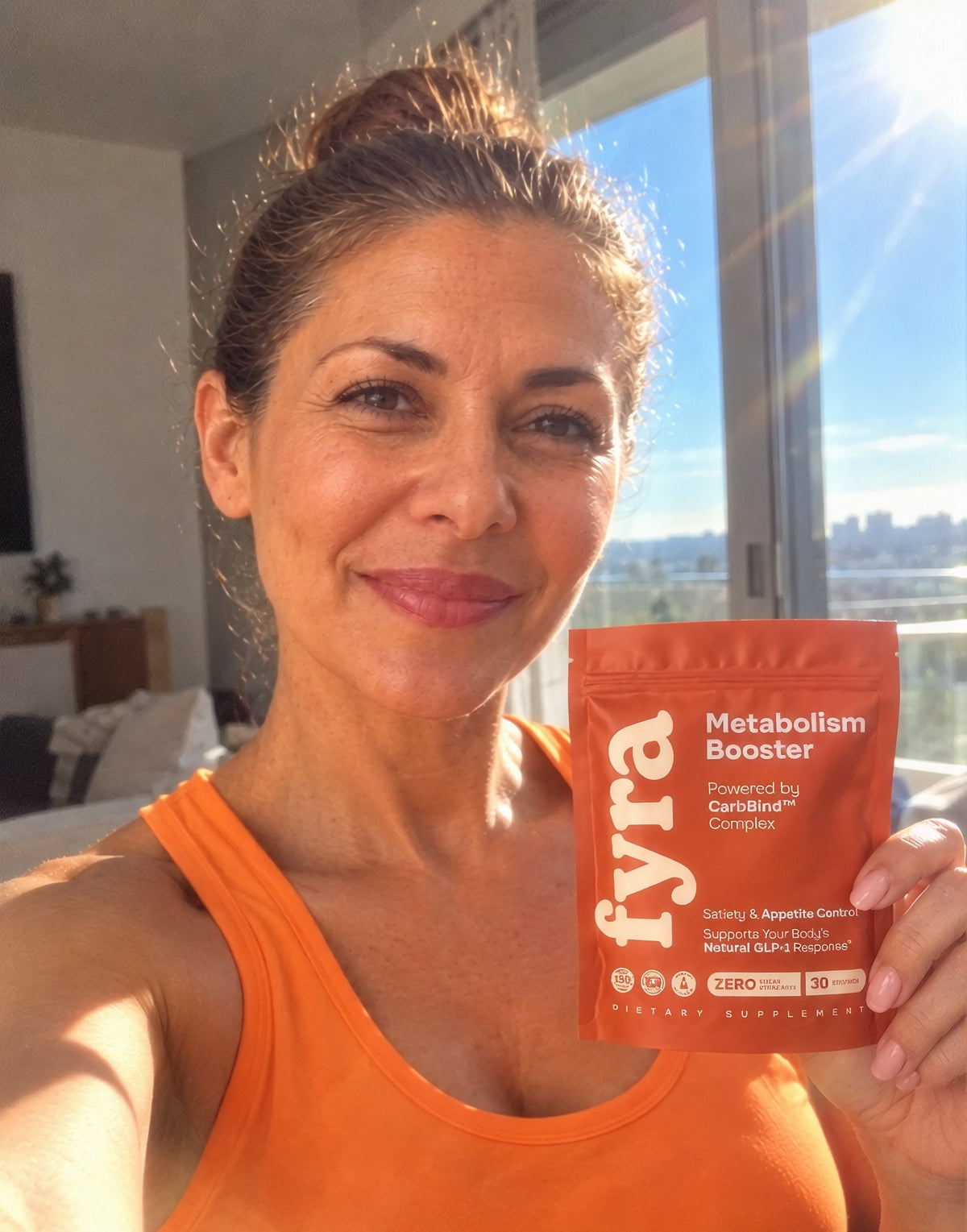 Woman in an orange tank top holding a pouch of FYRA Metabolism Booster